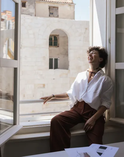 Person with curly hair wearing a white shirt and brown pants sitting on a windowsill laughing with sunlight coming through the open window.