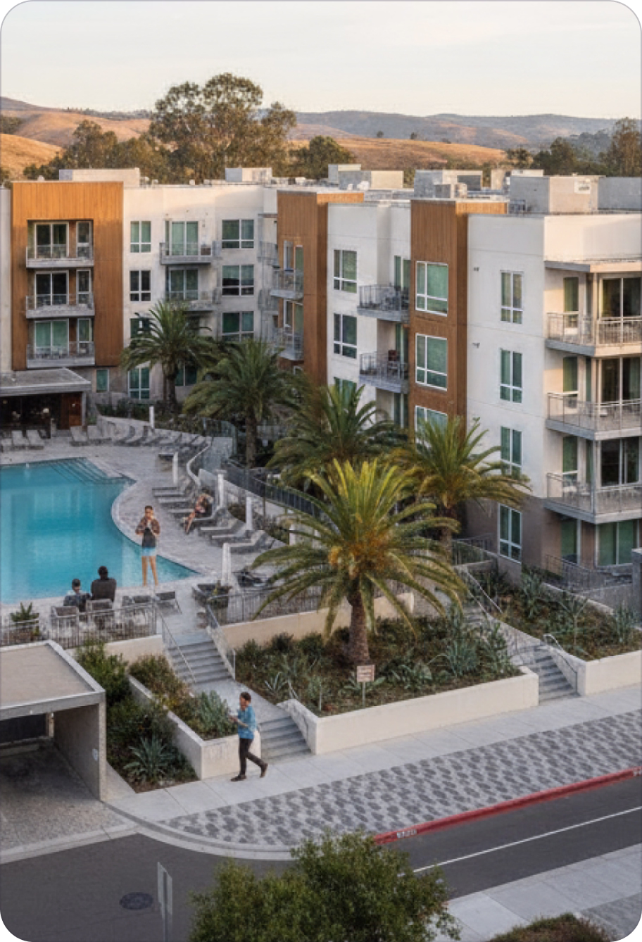 Modern apartment complex with balconies, palm trees, and a swimming pool with people relaxing nearby during daytime.