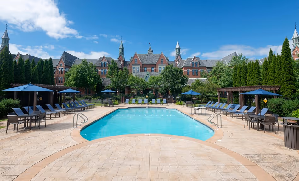 Outdoor swimming pool surrounded by lounge chairs with blue umbrellas in front of a large brick building with pointed towers and greenery.