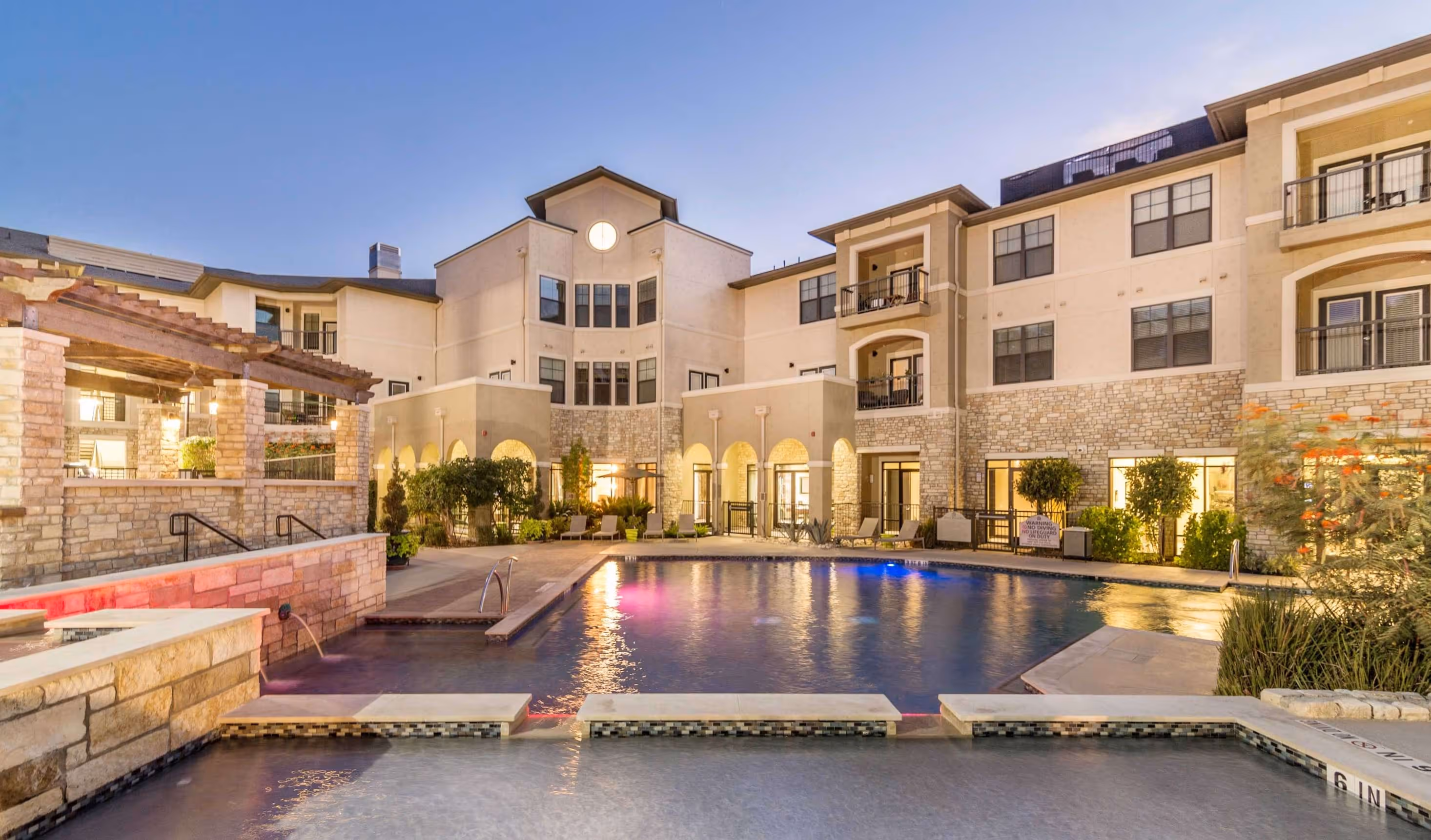 Evening view of an apartment complex courtyard with a lit swimming pool, lounge chairs, and stone and stucco buildings.