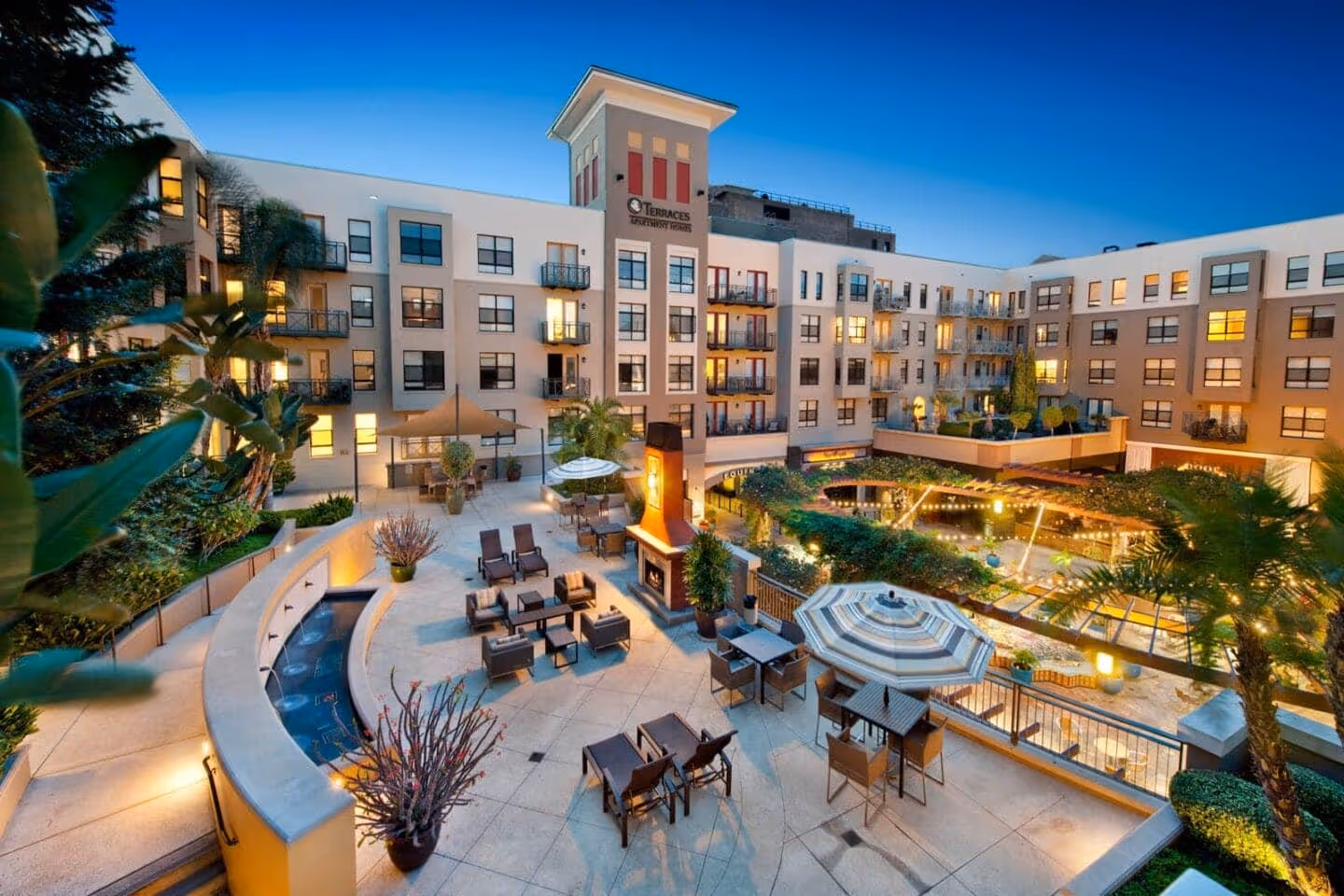 Evening view of a modern apartment complex courtyard with outdoor seating, umbrellas, a fireplace, and string lights.