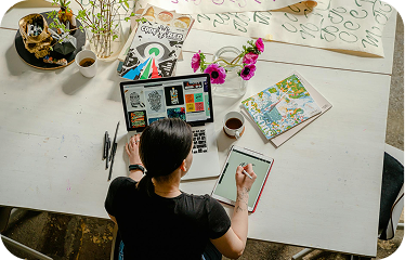 Person working on a laptop at a desk with pens, notebooks, flowers, and a cup of coffee nearby.