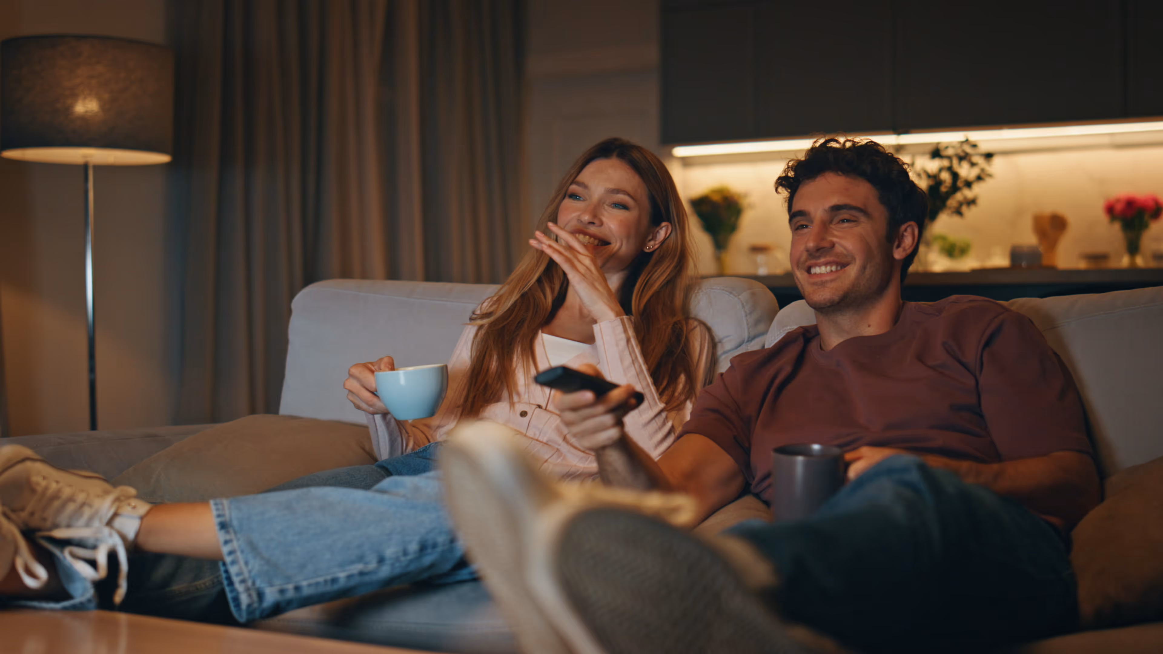 Smiling young couple sitting on a couch watching TV, holding mugs and a remote control in a cozy living room.