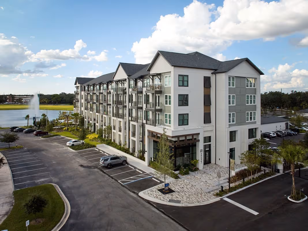 Modern four-story apartment building next to a parking lot and a lake with a water fountain under a partly cloudy sky.