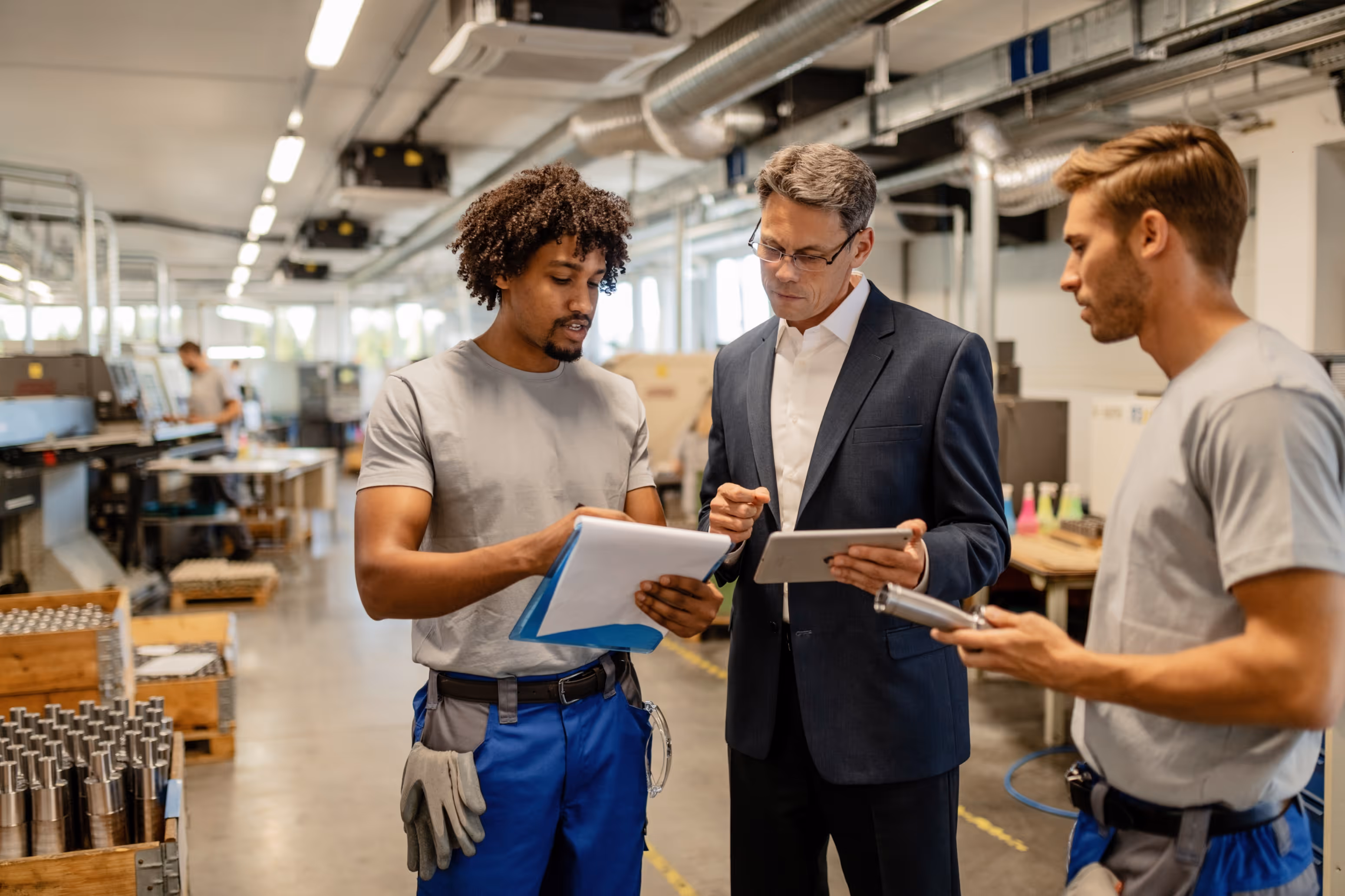 Two factory workers and a manager in business attire discussing documents and using a tablet in an industrial workspace.