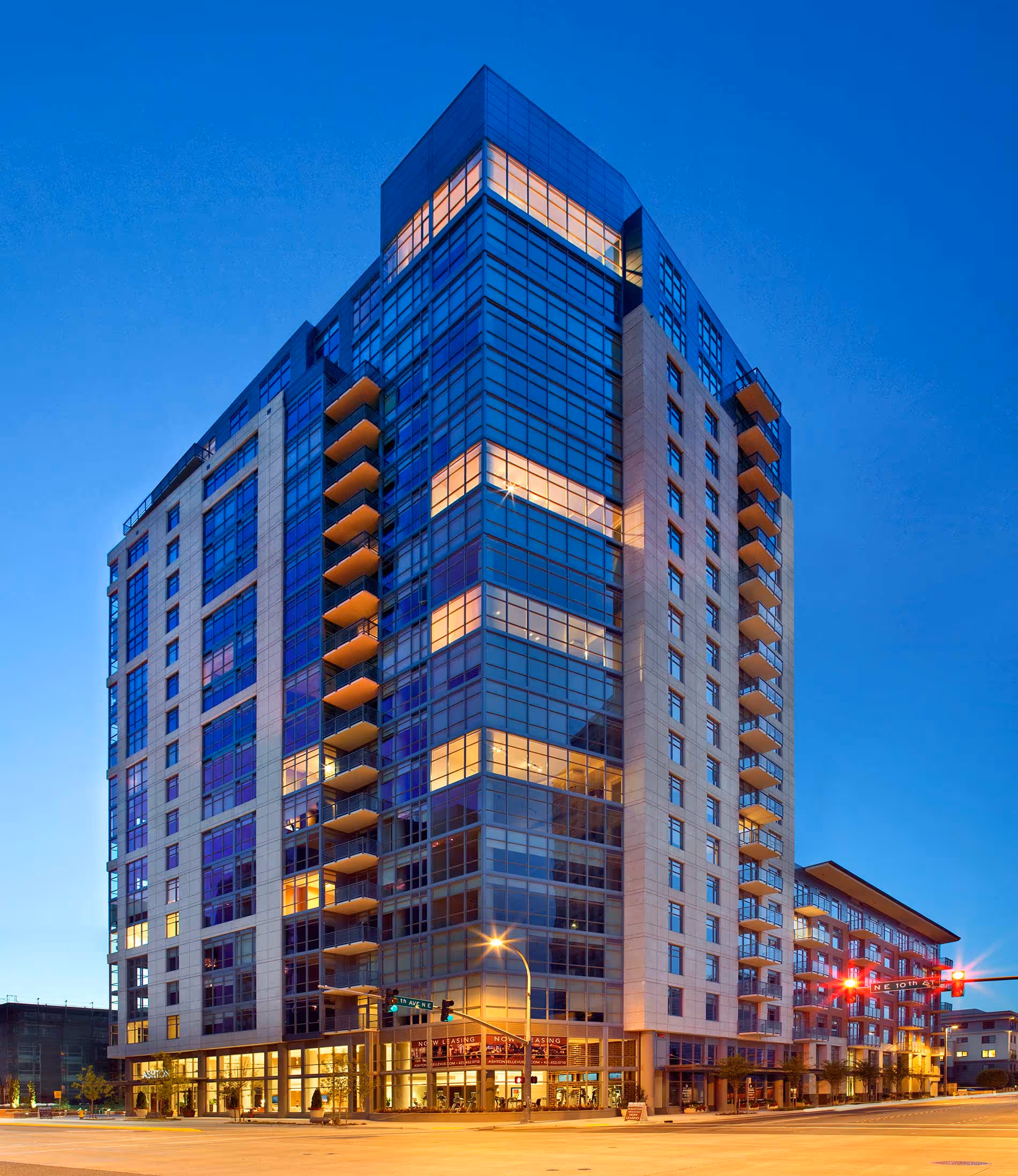 Modern multi-story residential building with glass windows and balconies illuminated at dusk on a city corner.