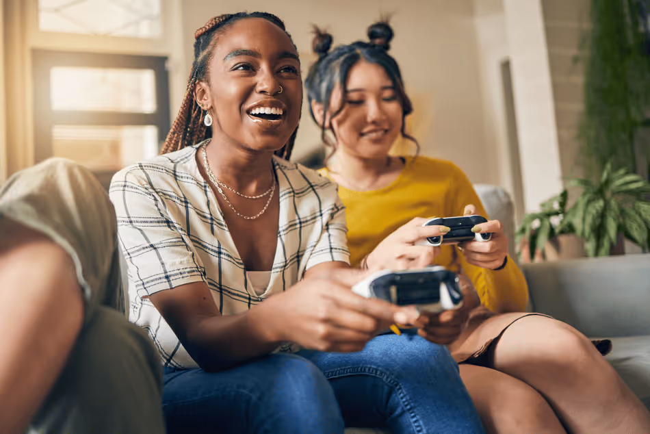 Two smiling female friends sitting on a couch holding video game controllers.