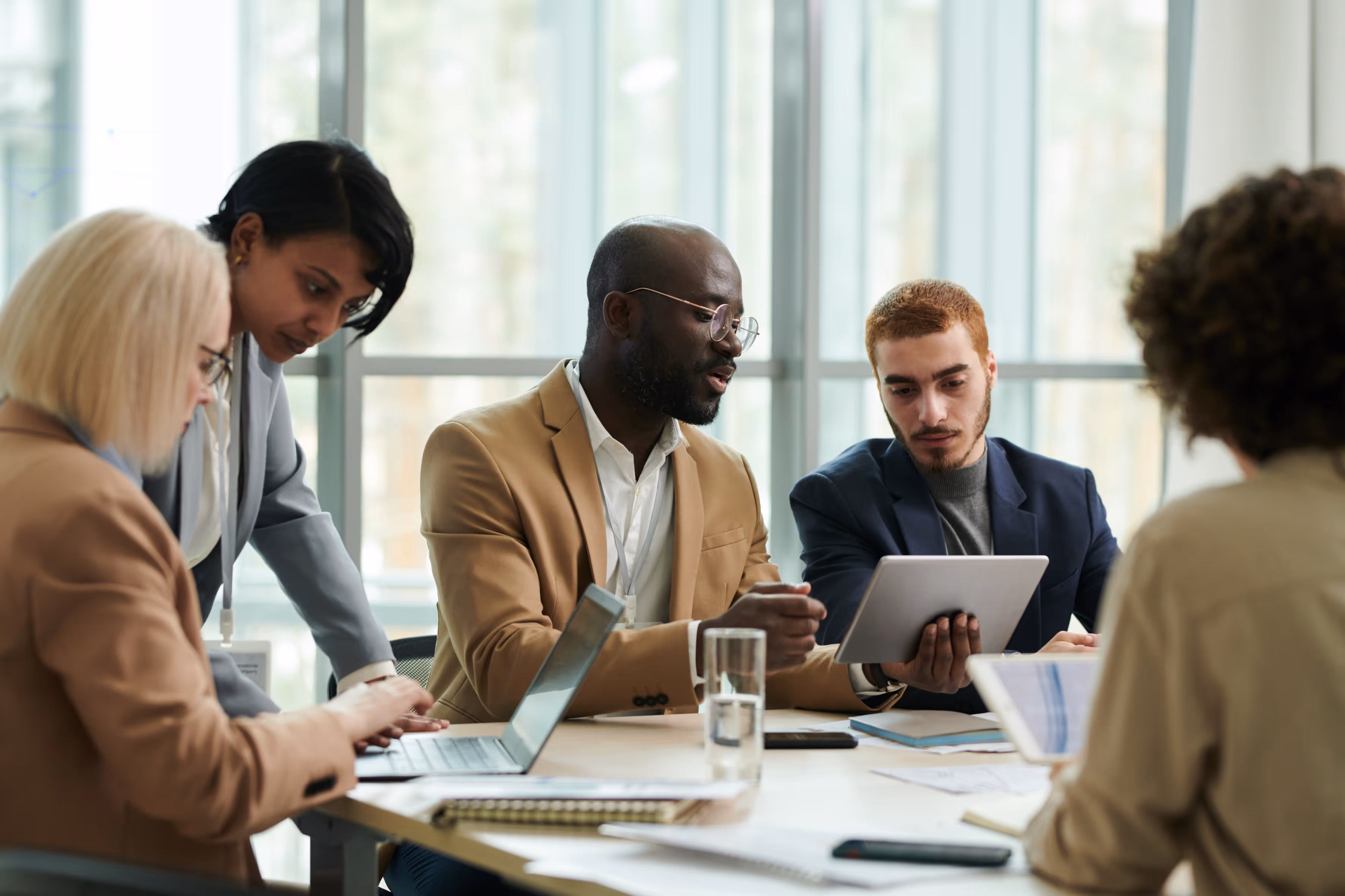 Five diverse professionals collaborating around a table with laptops and tablets in a bright office.