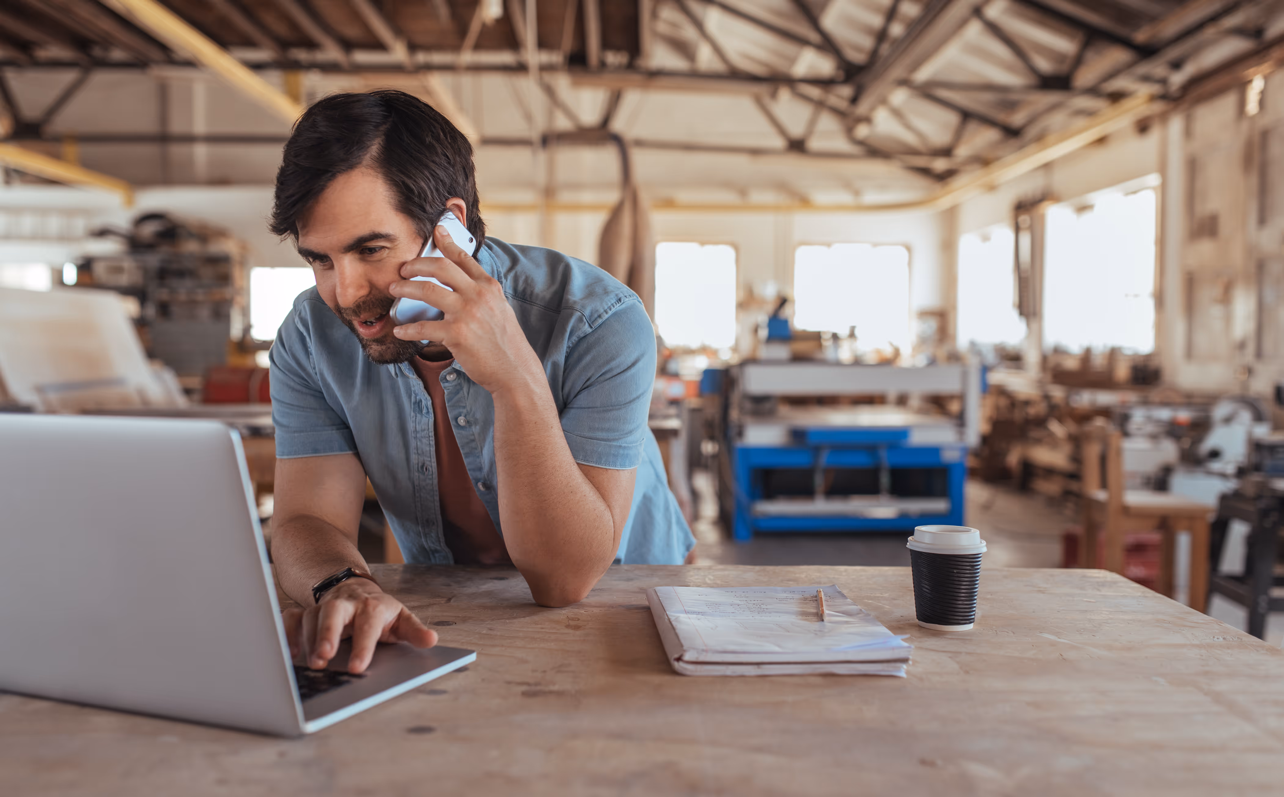 Man in a workshop talking on a smartphone and working on a laptop at a wooden table.
