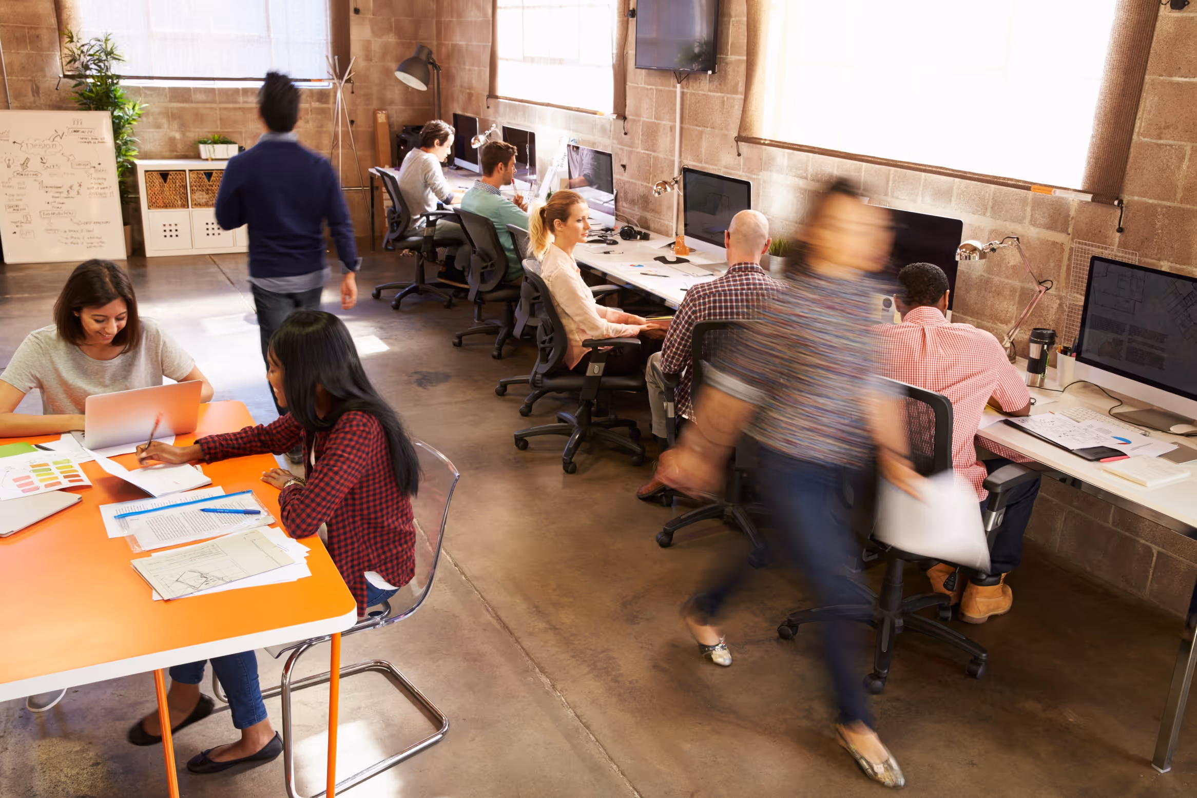 People working collaboratively and individually in a modern open office with computers, papers, and a whiteboard.