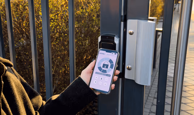 Person holding smartphone displaying a smart lock app near an electronic gate lock at sunset.