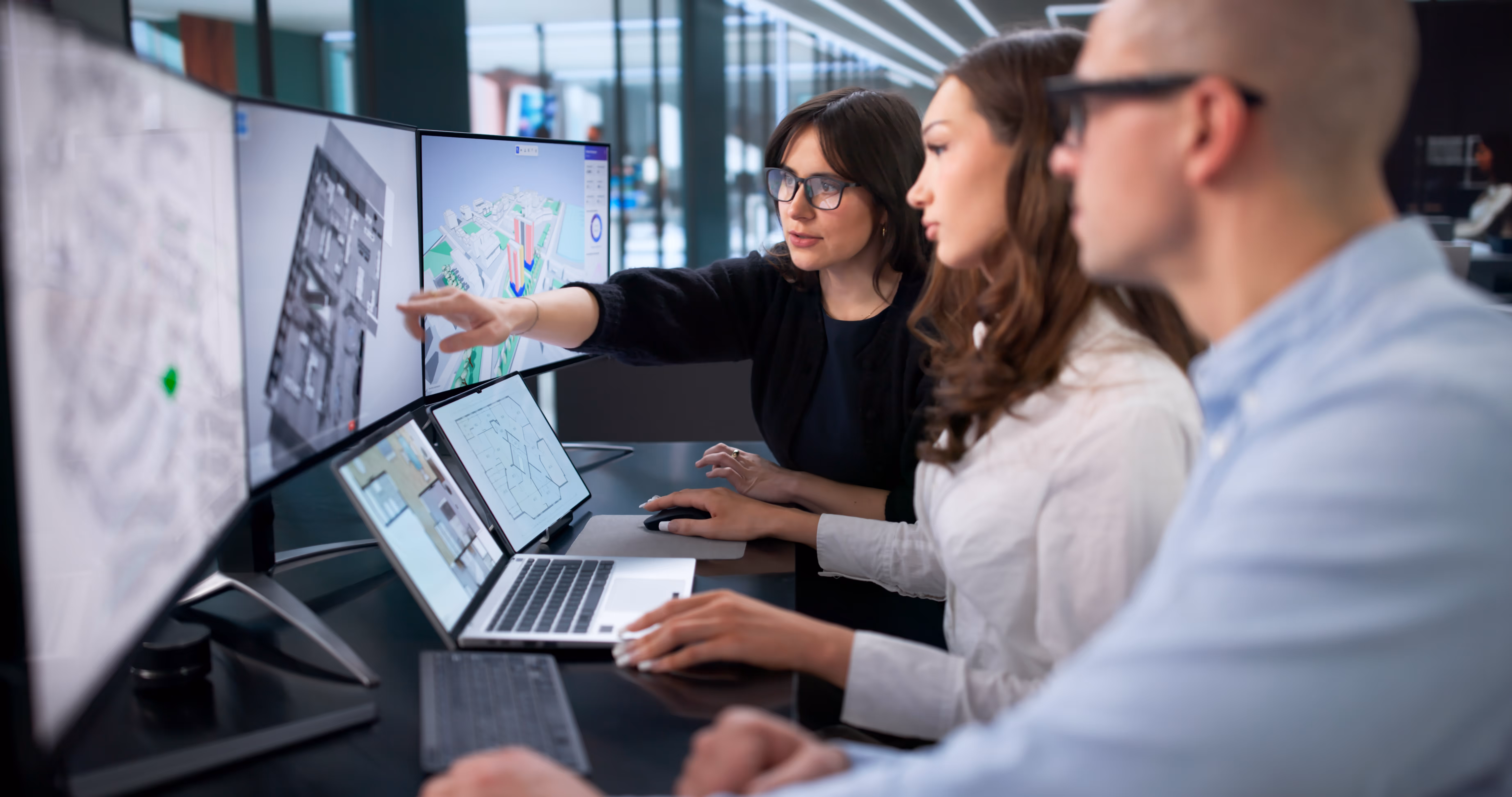 Three professionals reviewing architectural floor plans and city maps displayed on multiple screens and a laptop.