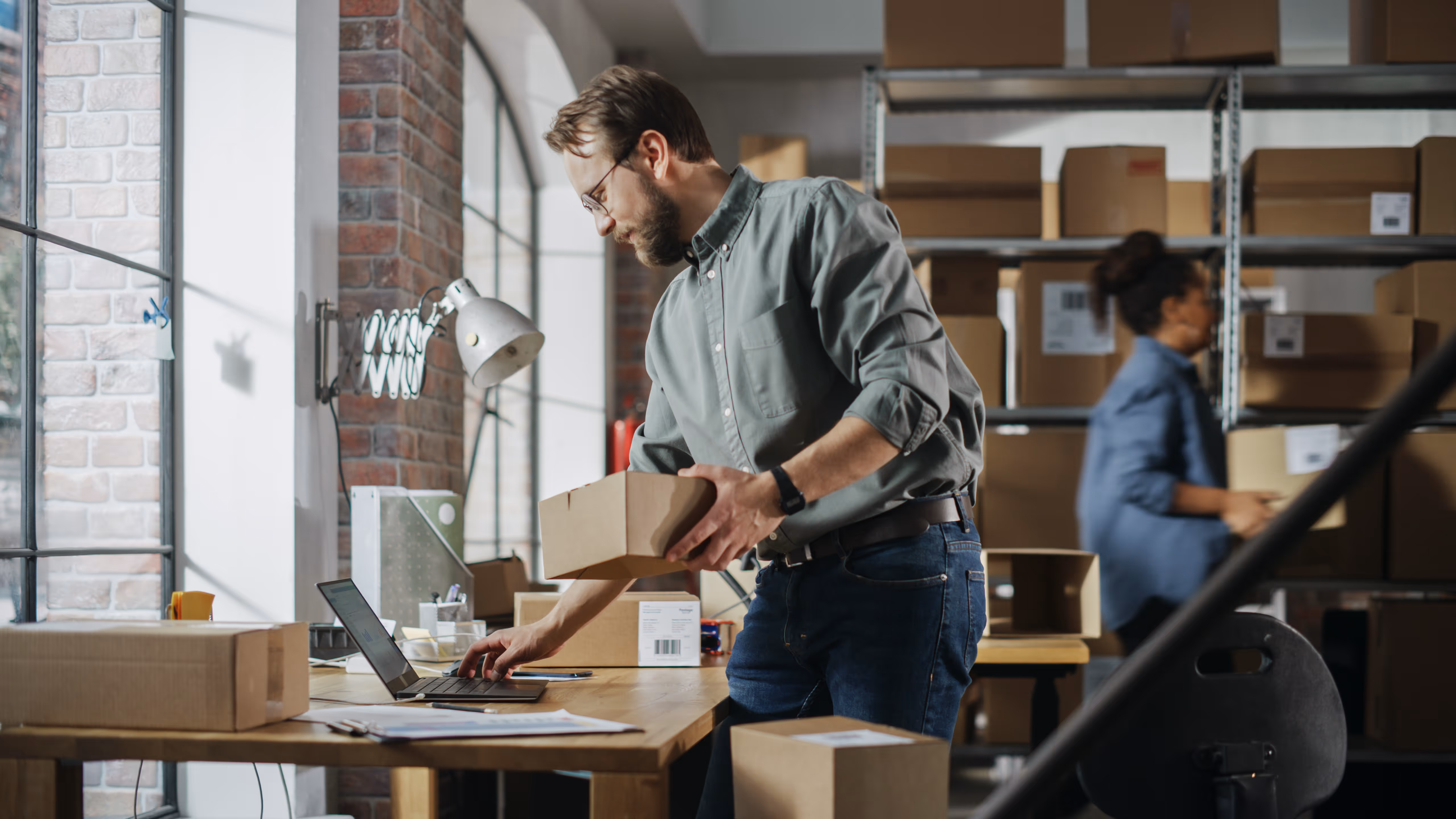 Man holding a cardboard box while working on a laptop in a room filled with stacked shipping boxes and shelves.