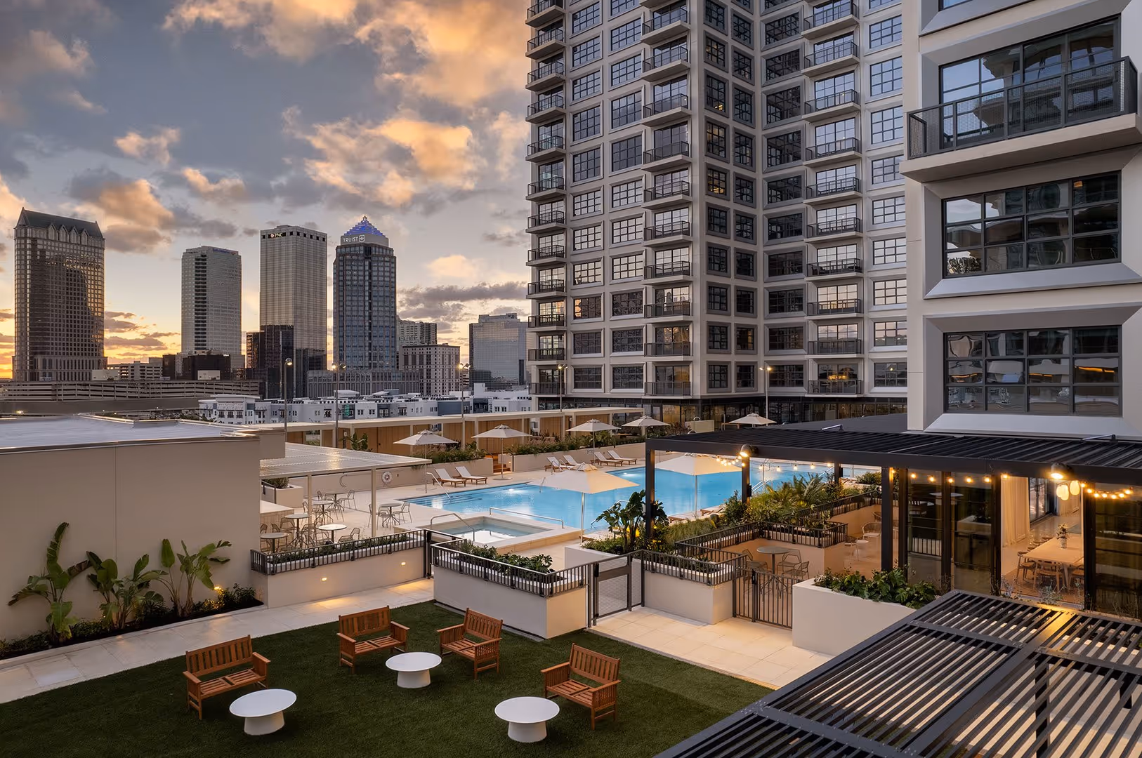 Apartment complex rooftop with swimming pool, lounge chairs, seating area, and city skyline at sunset.