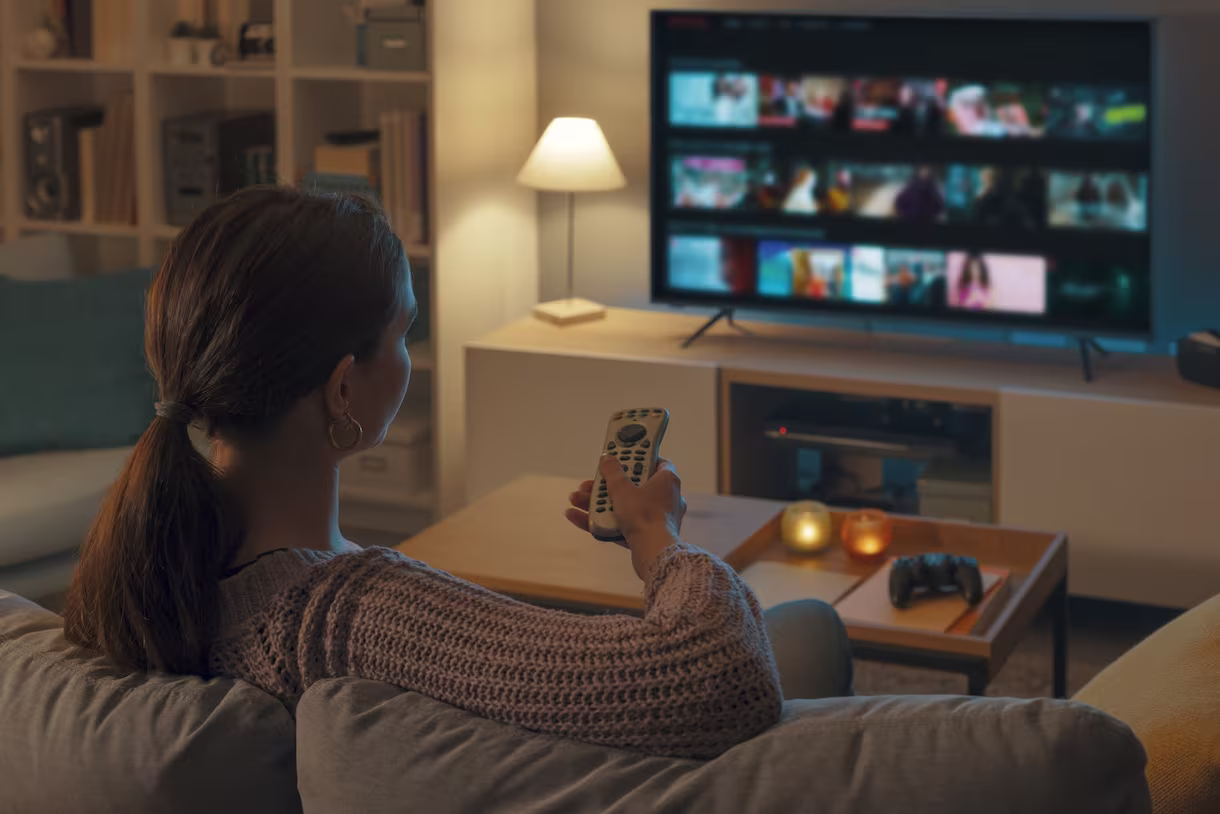 Woman sitting on a couch holding a remote control and watching a large TV screen with streaming content in a cozy living room.