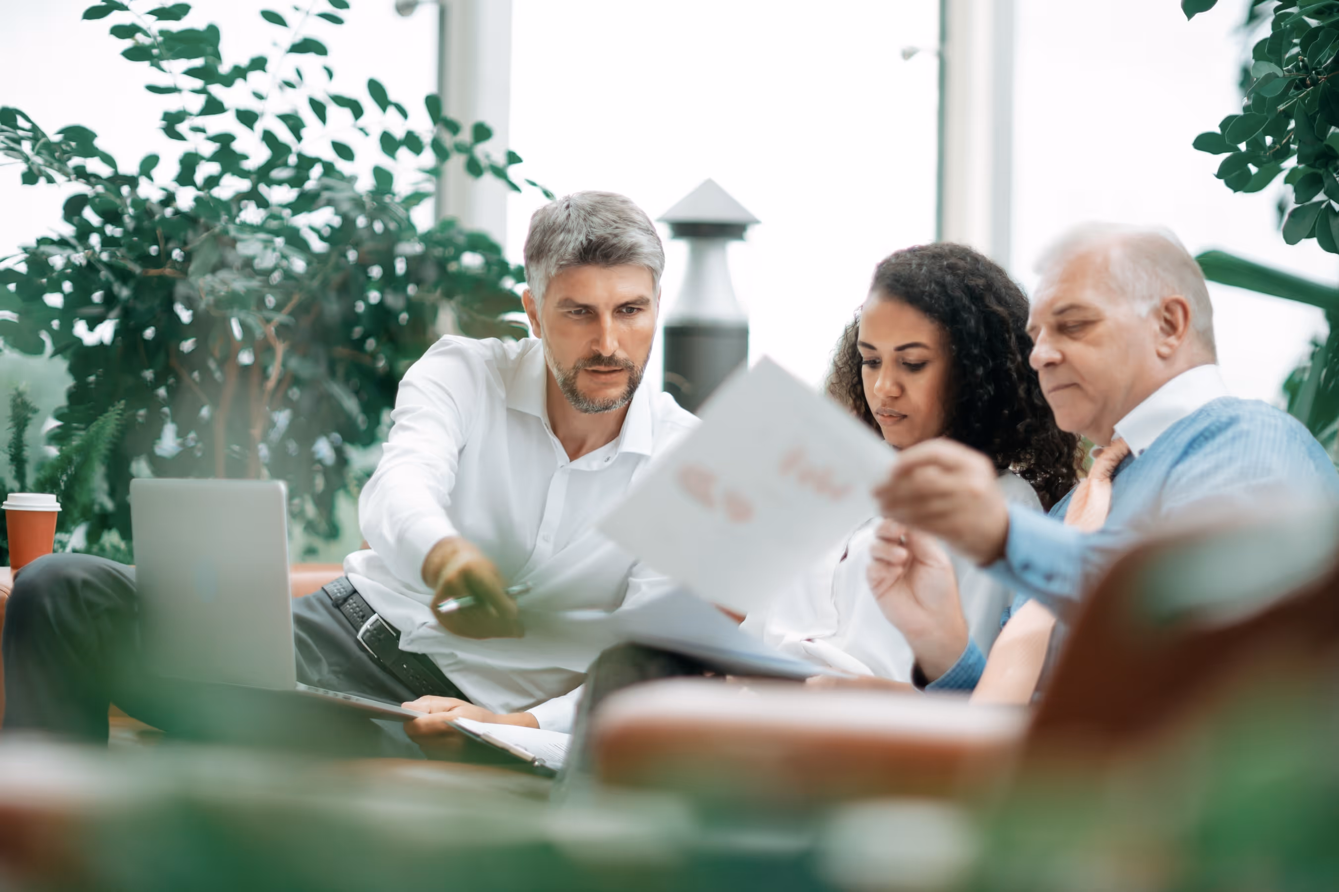 Three colleagues sitting on a couch in an office, reviewing documents together with a laptop nearby.