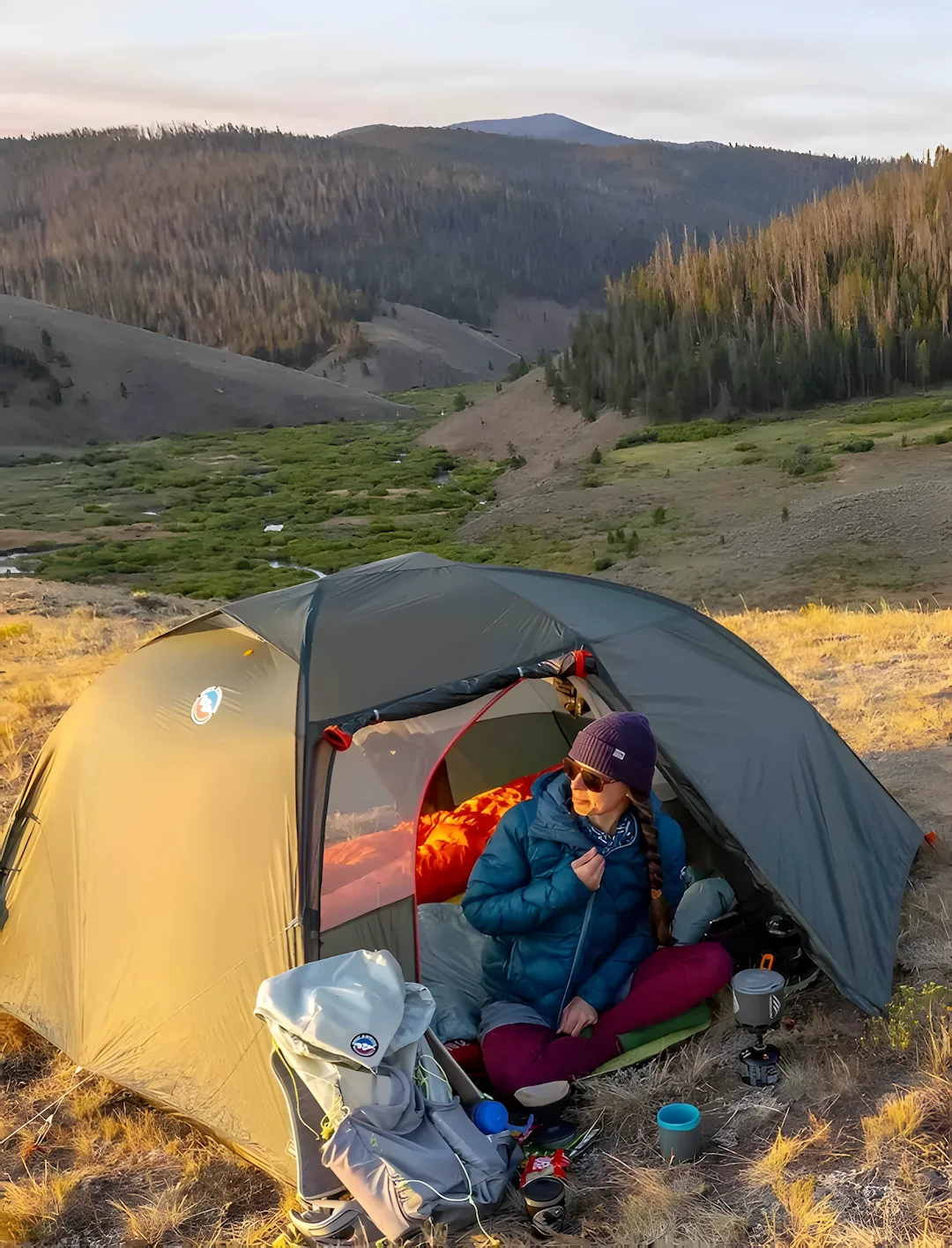 Femme dans une tente à la montagne, avec un sac à dos et un réchaud devant.