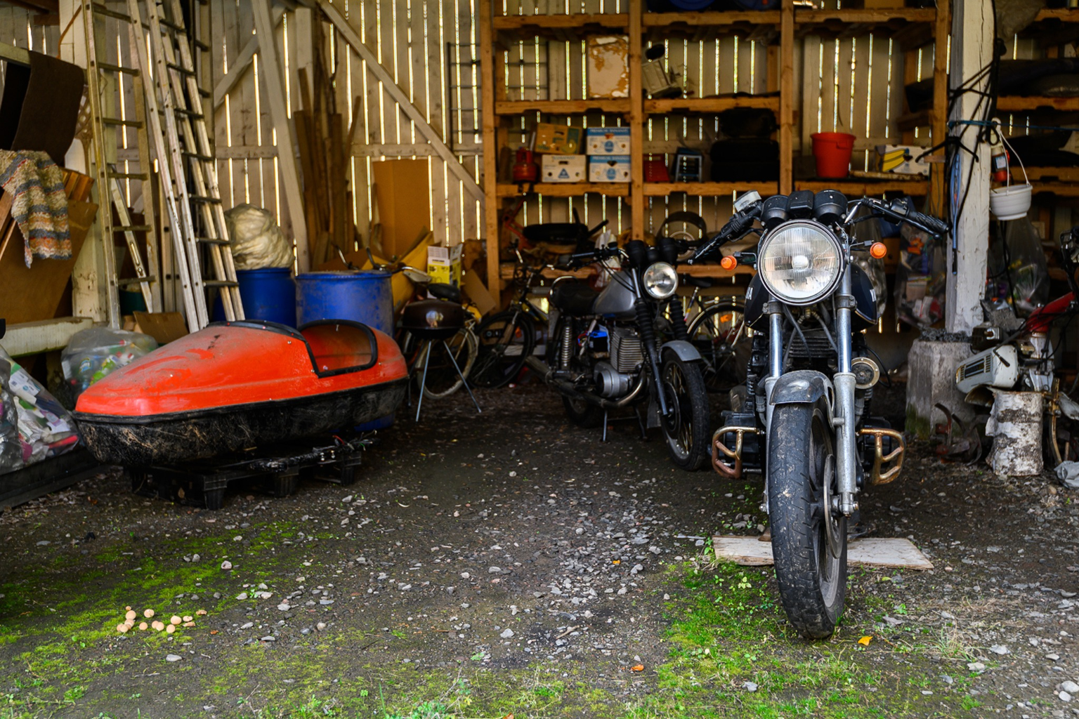 Zwei Motorräder und ein rotes Motorrad-Seitenwagen-Anhängergehäuse in einer Scheune mit Regalen und Leitern.