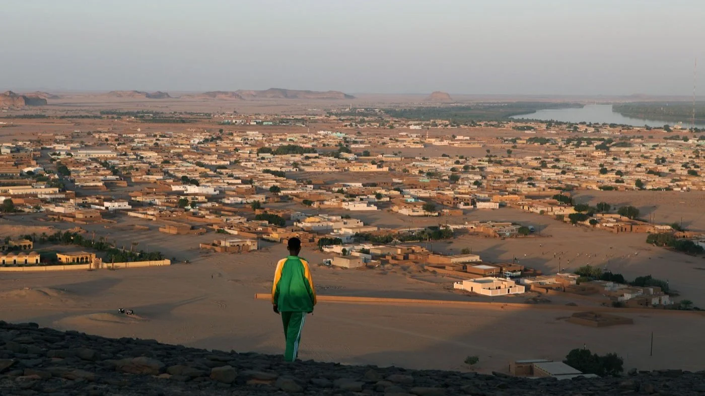Person in green and yellow tracksuit standing on a hill overlooking a sprawling desert town with numerous flat-roofed buildings and a river in the distance.