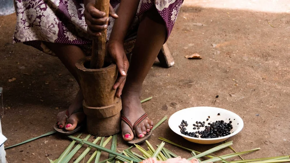 Person using a wooden mortar and pestle to grind food with a bowl of black beans nearby on the ground.