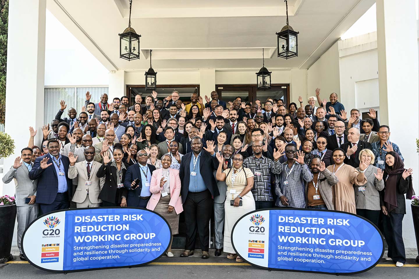 Large diverse group of people posing and waving in front of a building with two signs reading 'Disaster Risk Reduction Working Group' for G20 South Africa 2025.