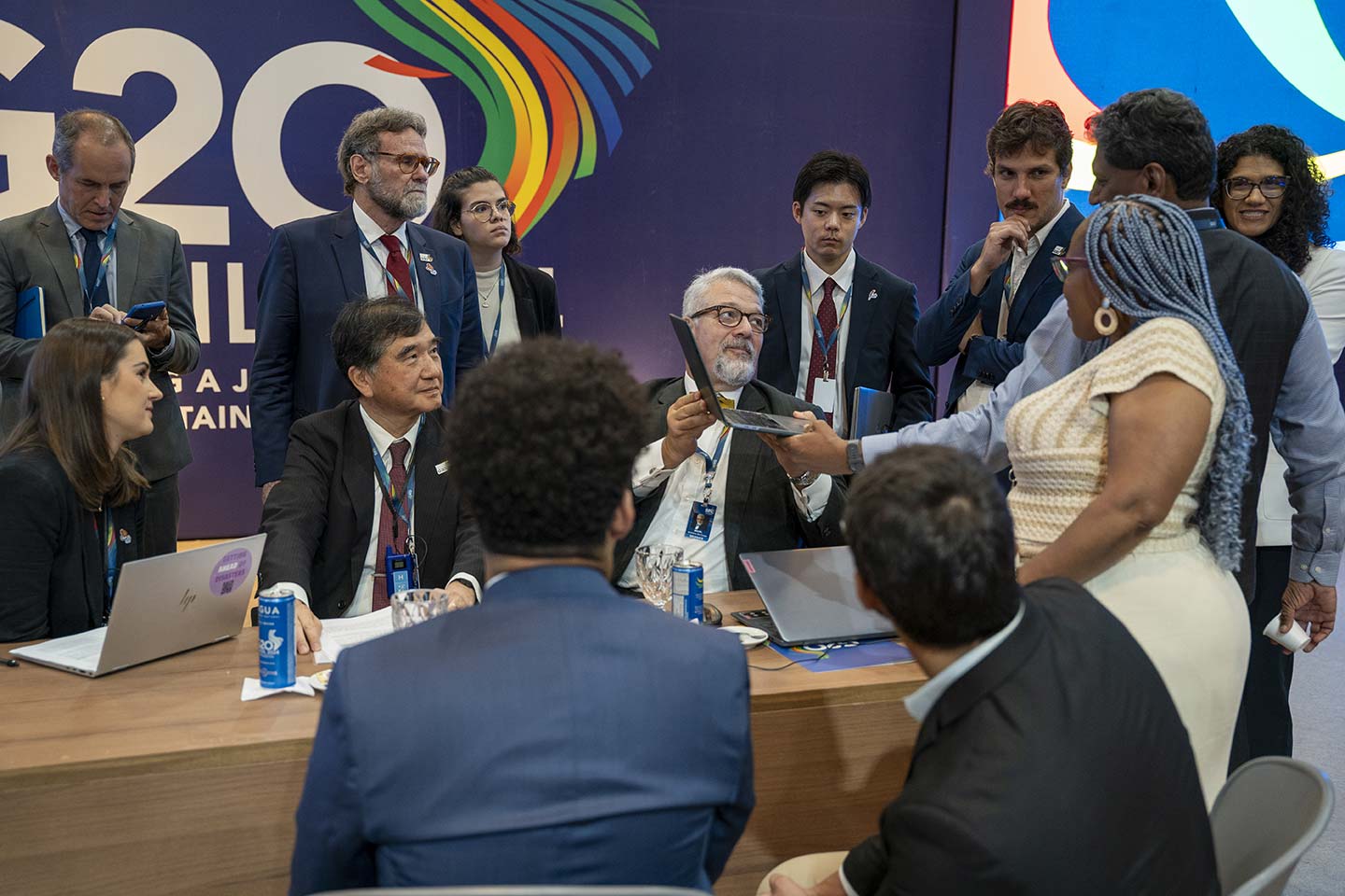 Group of diverse professionals in formal attire gathered around a table at a G20 meeting, one man holding up a laptop for discussion.