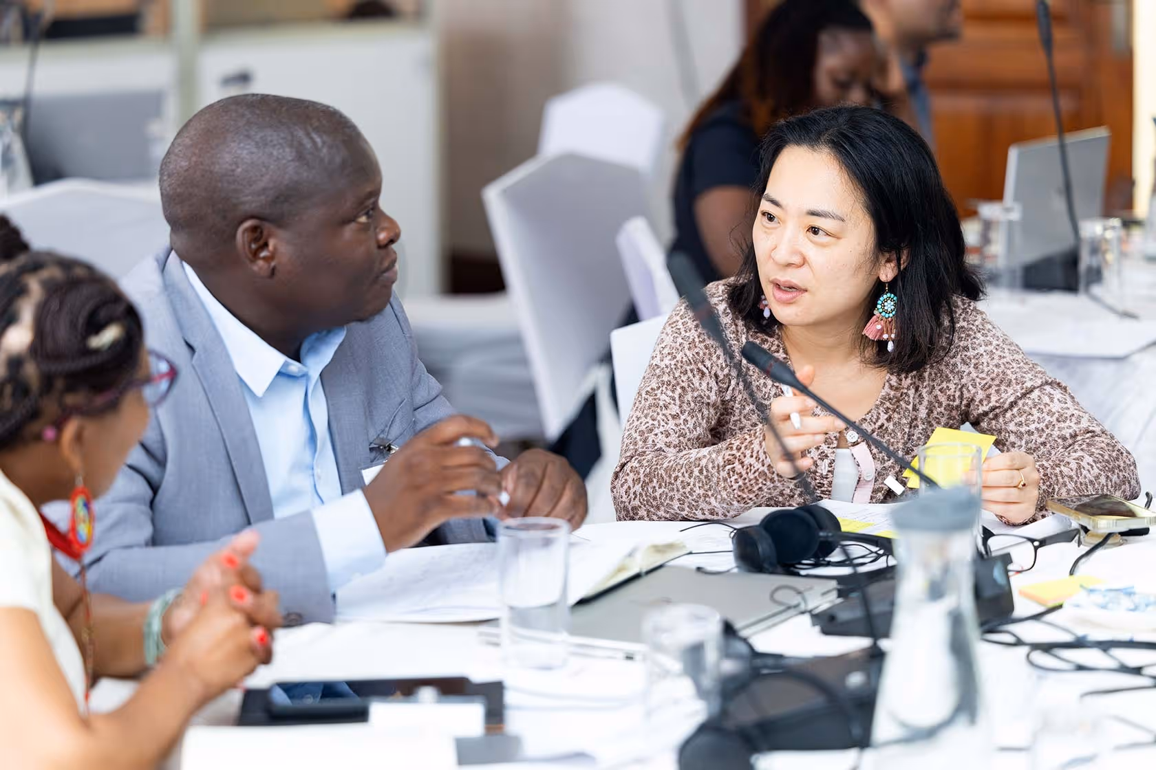 Three people engaged in a discussion around a table with papers, laptops, and microphones.