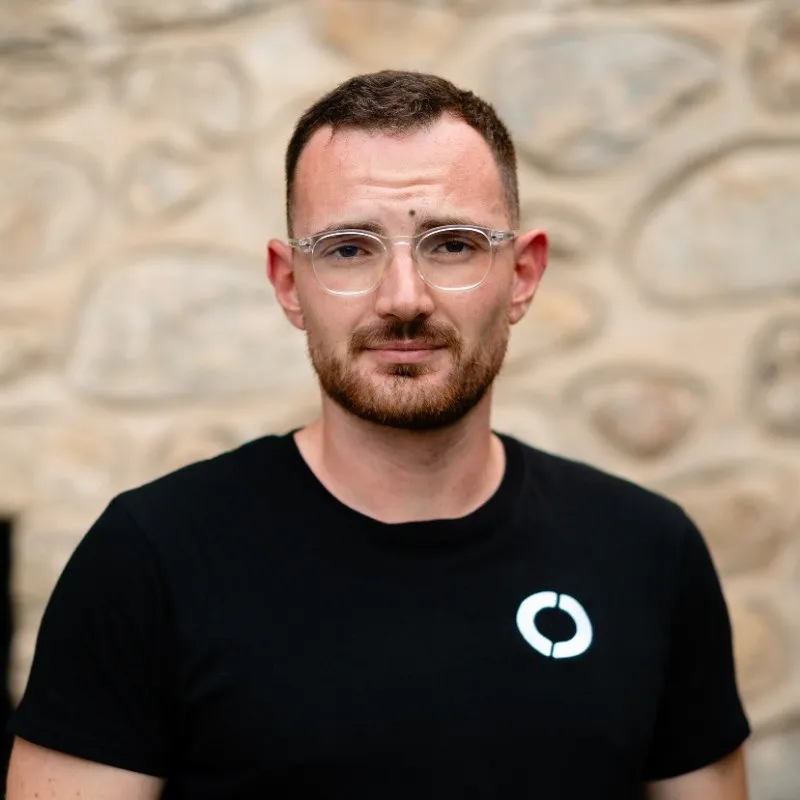 Man with short dark hair, beard, and clear glasses wearing a black t-shirt with a white circular logo, standing in front of a stone wall.