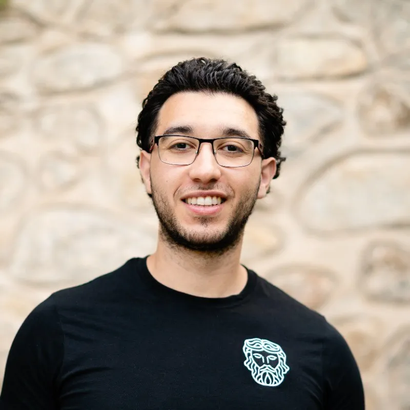 Smiling man with glasses and curly black hair wearing a black shirt with a white bearded face logo, standing in front of a stone wall.