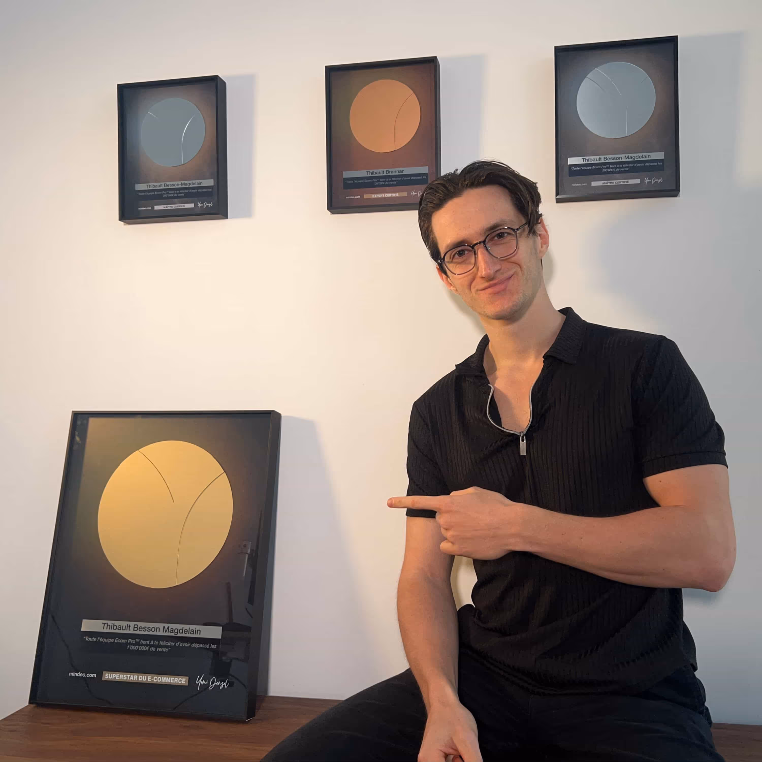 Man wearing glasses and a black shirt sitting and pointing to a gold e-commerce award plaque on a table, with three framed awards hanging on the wall behind him.