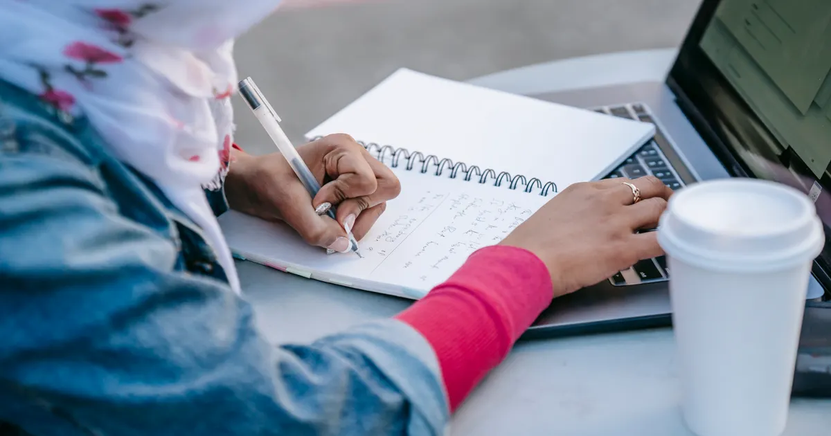 A student writing a blog post on a laptop next to an open notebook