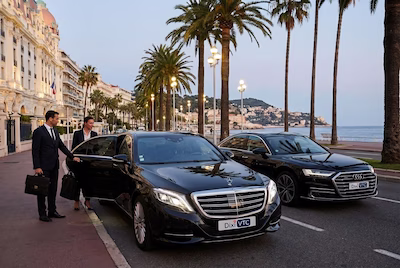 Chauffeur ouvrant la porte d’une Mercedes Classe S sur la Croisette à Cannes, avec les palmiers et la mer en arrière-plan.
