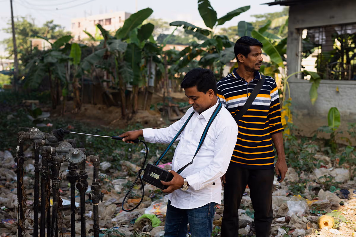 Two men inspect an outdoor water pipe system in a cluttered area with green plants and a rundown building in the background.