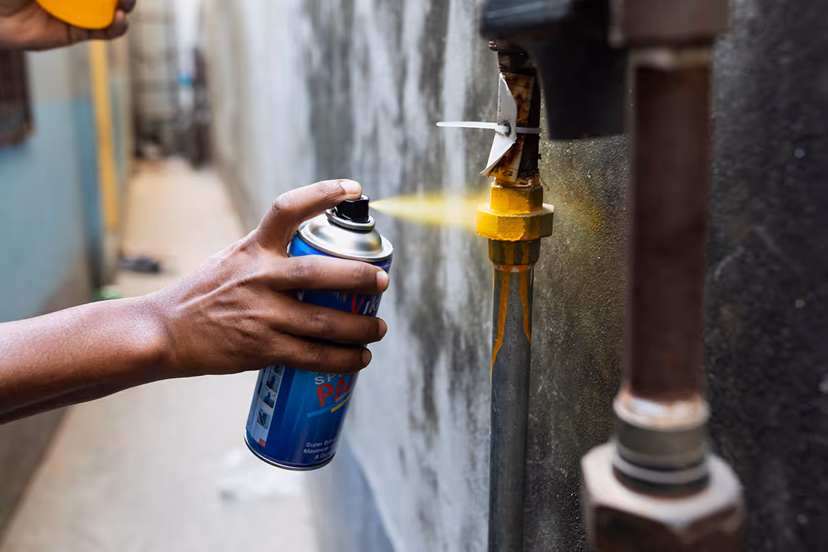 Hand spraying lubricant spray on a yellow valve attached to a pipe on a wall.