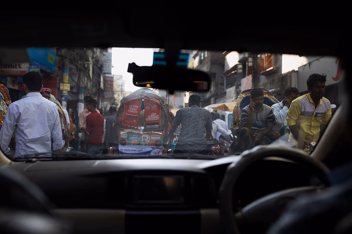 View through a car windshield of a busy street crowded with pedestrians and rickshaws in an urban area.