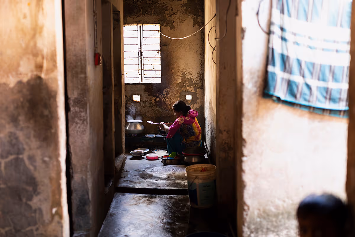 A person sits on the floor in a narrow, dimly lit kitchen stirring a pot on a stove with steam rising.