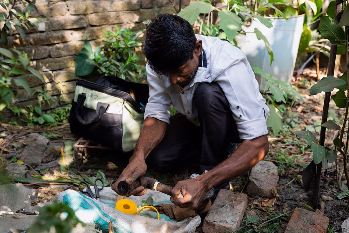Man kneeling outdoors repairing a rusty metal pipe with tools and a bag nearby.