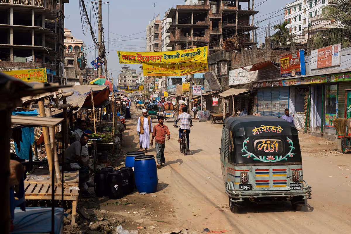Busy street in a city with pedestrians, a person riding a bicycle, a tuk-tuk, market stalls, and unfinished buildings under a clear sky.
