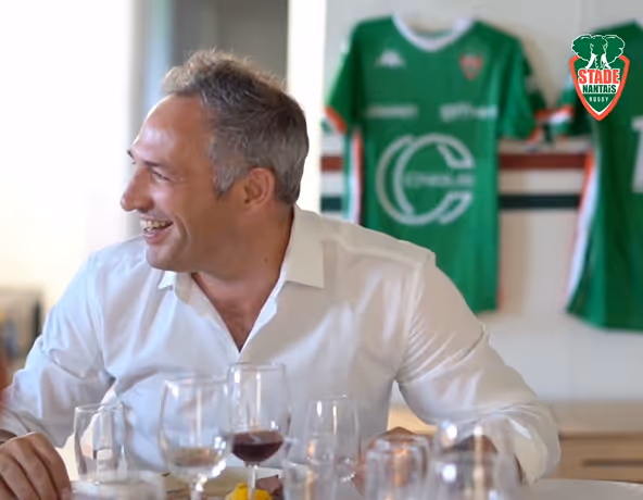 Homme souriant dans les vestiaires du Stade Nantais, avec un maillot en arrière-plan.