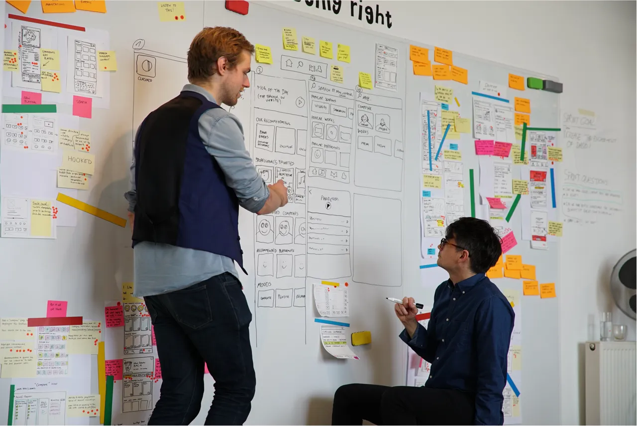 AI Workshops- Two men collaborating in an office, discussing wireframes and sticky notes on a whiteboard wall.