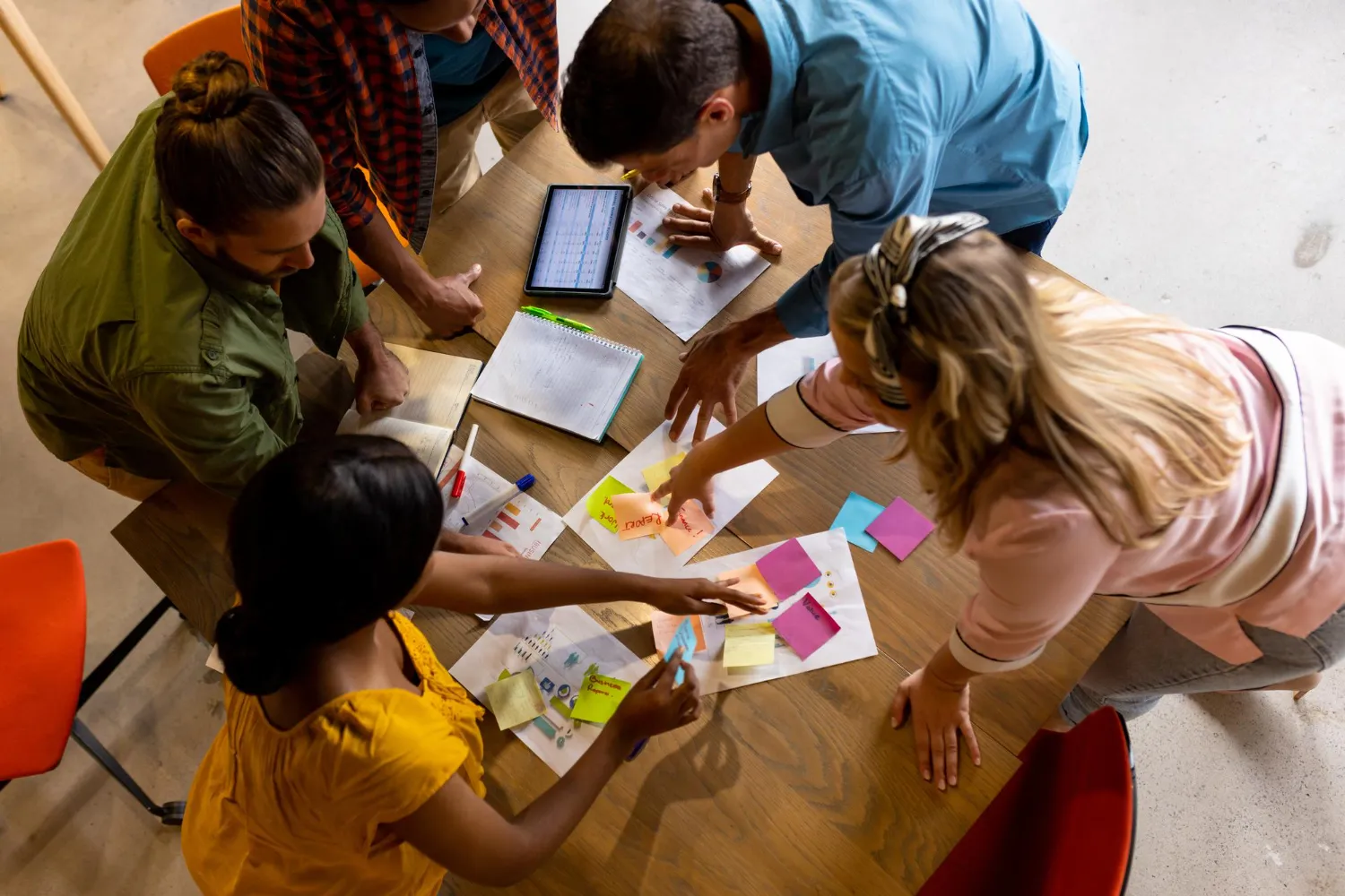 Five diverse people brainstorming around a table covered with sticky notes, papers, and a tablet displaying charts.