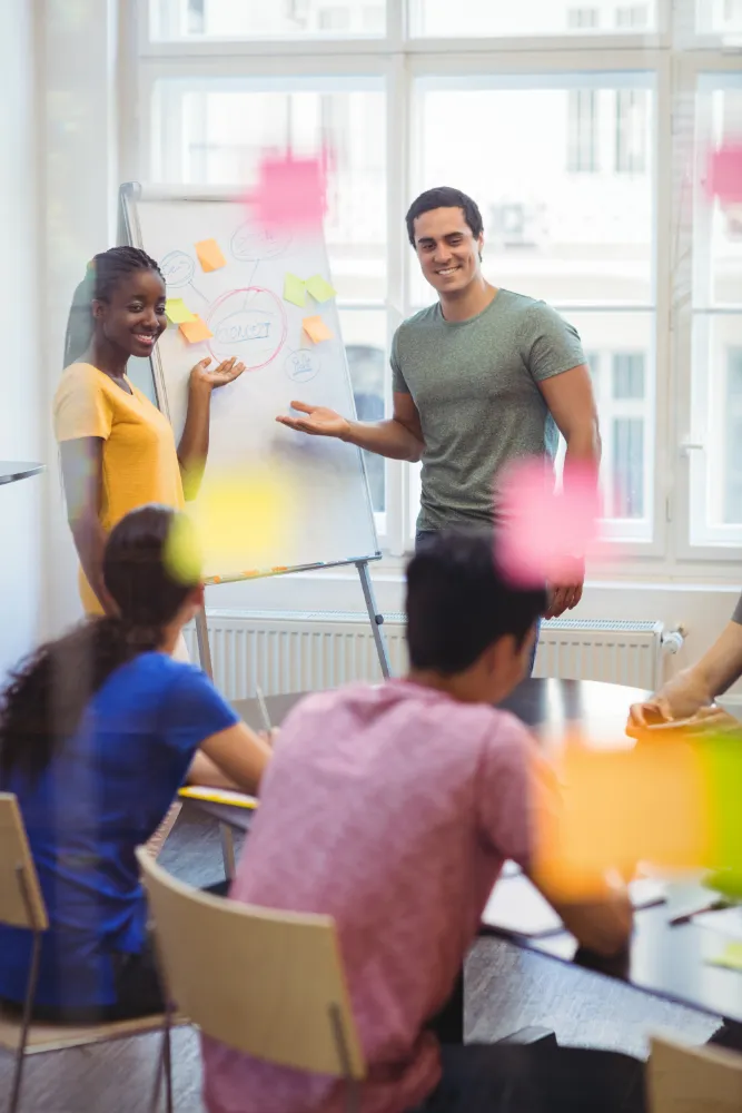 Two people smiling and presenting ideas on a flip chart to a seated group in a bright meeting room with sticky notes on the glass.