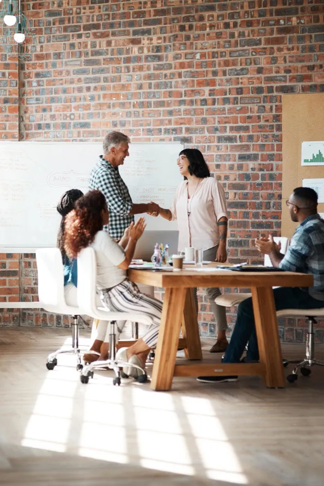 Two colleagues shaking hands and smiling in an office while others at a table applaud.
