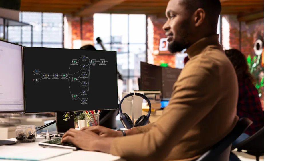 Man in a brown turtleneck working at a computer with a flowchart displayed on the monitor in a bright modern office.
