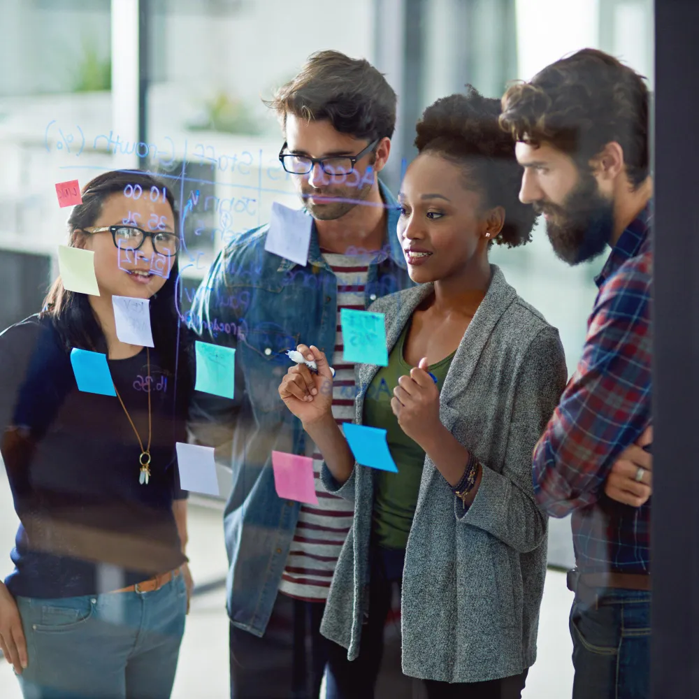 Four young professionals discussing notes on a glass wall covered with colorful sticky notes and handwritten text.