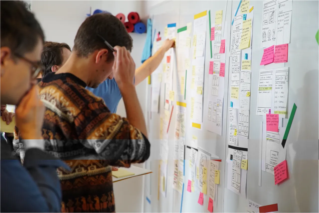 Group of people analyzing and discussing notes and sketches on a whiteboard covered with colorful sticky notes and papers.
