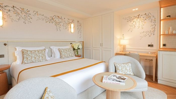 Chambre d'hôtel lumineuse avec un grand lit double, deux fauteuils gris autour d'une petite table ronde, décor mural floral et bureau avec lampe.
