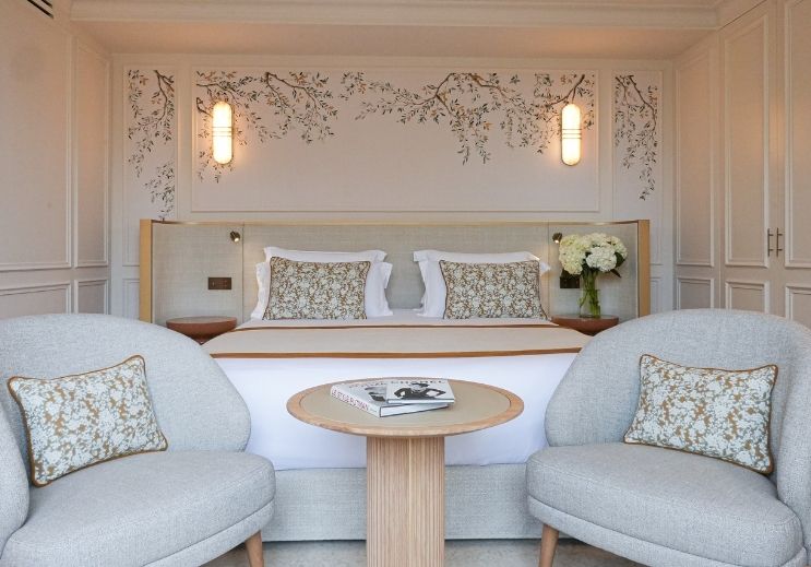 Chambre élégante avec un grand lit double, deux fauteuils gris avec coussins à motif floral, table ronde en bois clair et bouquet de fleurs blanches.