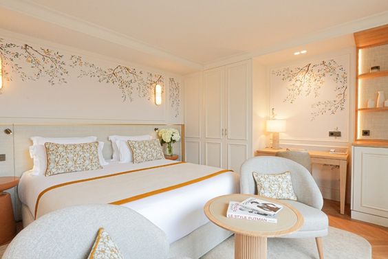 Chambre lumineuse avec lit double, fauteuils gris, table ronde en bois, décor floral peint sur les murs, et mobilier blanc élégant.