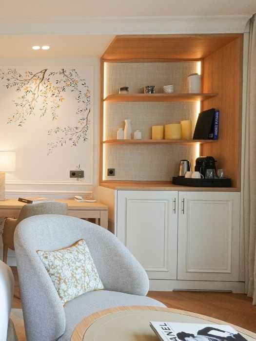 Coin salon avec fauteuil gris clair, coussin décoratif, table ronde avec magazine, étagères en bois intégrées avec bougies et cafetière, et décoration murale feuillue.